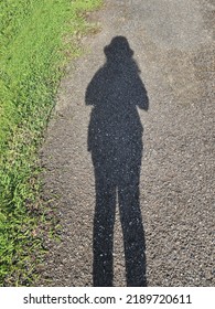 Looking down at the shadow of a woman on a concrete road, to the left is a green lawn. In shadow she was wearing a hat and can see long legs. Sunlight shines through the body, creating funny shadows.