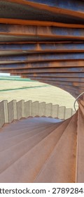 Looking down a spiral staircase inside a Corten steel architectural sculpture near Ribe, Denmark. Weathered rusted steel layers arch overhead, curved steps descend below. Agricultural fields visible through the opening.
