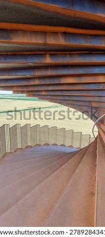 Looking down a spiral staircase inside a Corten steel architectural sculpture near Ribe, Denmark. Weathered rusted steel layers arch overhead, curved steps descend below. Agricultural fields visible through the opening.