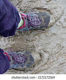 Looking Down At Two Feet In Pink And Grey Walking Boots That Are Sinking Into Thick, Sticky Clay Mud. Blue Trousers Have Been Tucked Into Green Socks. The Approach Shoe Laces Are Purple - Mauve Grey.