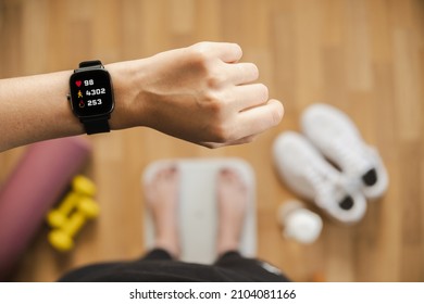 Looking at fitness tracker displaying heart rate, steps count and burnt calories while standing on scales, first person view. Smart watch on female hand, point of view, concept of staying fit at home