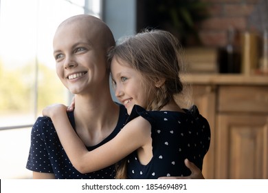 Looking In Future With Hope. Young Hairless Mother With Cancer Diagnosis Standing By Wide Window At Home Embracing Beloved Little Daughter Looking At Distance With Smile Optimism Belief In Recovery