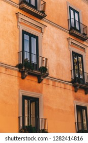 Looking Up From Ground Level At A Group Of Italian Apartment Balcony Windows And Plants With An Orange Terracotta Facade And One Window Lit Up.