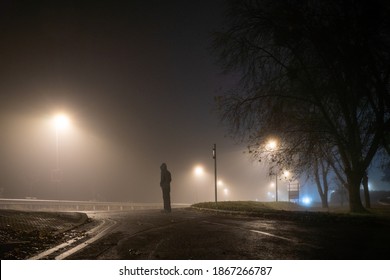 Looking Up At A Hooded Figure Standing On A Road With Street Lights On An Atmospheric Foggy Winters Night. UK.