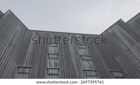 Looking up at the imposing and geometric facade of a bleak, weathered building. The monochromatic tones and stark lines create a somber, industrial atmosphere.