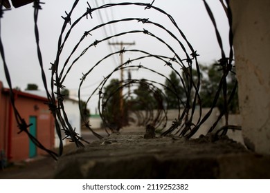 Looking Inside A Barbed Wire Fence On Top Of A Rock Wall. It's Cloudy, There Are Telephone Poles And A Small Red House In The Background.