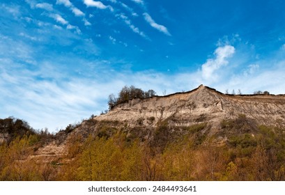 Looking up at a jagged eroded cliff face under a beautiful cloudy blue sky with many trees and plants below. - Powered by Shutterstock