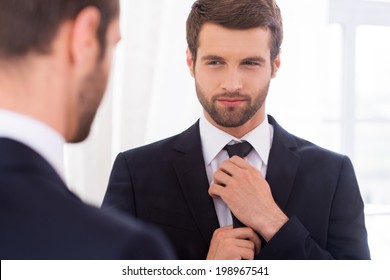 Looking just perfect. Handsome young man in formalwear adjusting his necktie and smiling while standing against mirror - Powered by Shutterstock