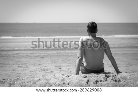 looking at a man his back on the beach he is looking and relaxing at the seaside in retro old fashioned style. There is a heart painted on him with suncream in black and white