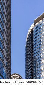 Looking up at a narrow vertical space between two towering curved glass skyscrapers framing the blue sky. The scene represents urban density, corporate architecture, and dramatic city perspectives.