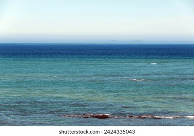 Looking out over a calm, flat sea in different shades of blue, towards a hazy horizon with mountains in the distance.