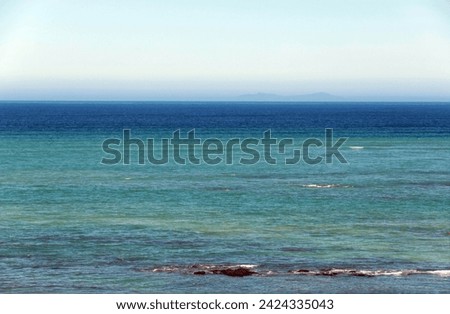 Looking out over a calm, flat sea in different shades of blue, towards a hazy horizon with mountains in the distance.