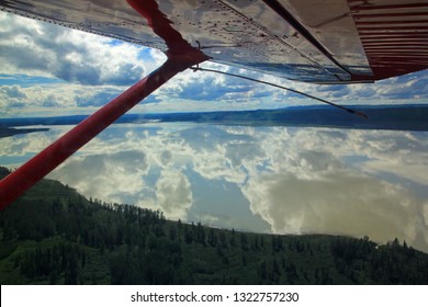 Looking Out A Small Plane Window To See The Clouds Reflecting In A Calm Lake Below