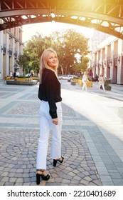 Looking Over Her Shoulder Attractive Young Blond Woman Walking Down The Road, Looking At The Camera. She Is Walking On A Pavement On A Sunlit Street.