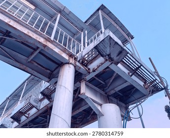 Looking up photograph capturing the complex, weathered steel structure of an elevated pedestrian bridge or skywalk. The white-painted beams, girders, and large cylindrical support pillars show significant rust, corrosion, and wear, giving the structure a rugged, industrial character.