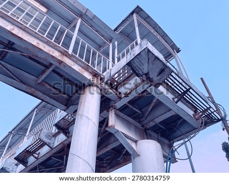 Looking up photograph capturing the complex, weathered steel structure of an elevated pedestrian bridge or skywalk. The white-painted beams, girders, and large cylindrical support pillars show significant rust, corrosion, and wear, giving the structure a rugged, industrial character.