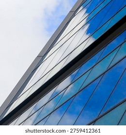 Looking up at the sharp diagonal edge of a modern glass building splitting the frame between muted overcast sky and vivid blue cloud reflections in the facade. Converging lines and repeating glass panels create dynamic upward perspective. Bold contrast between the grey sky and the saturated blue reflections gives the building a surreal, atmospheric quality.