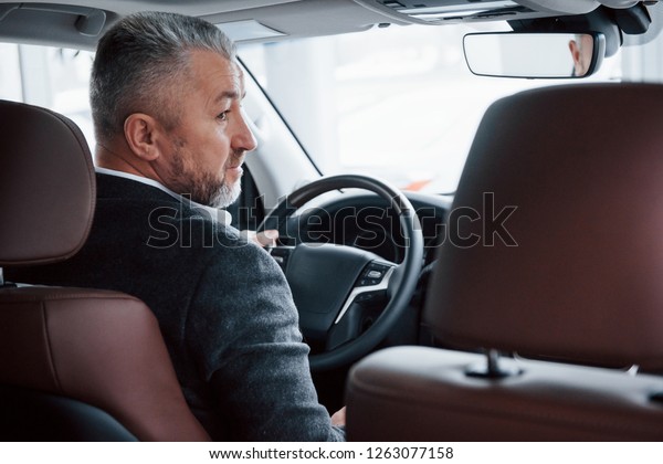 Looking to the side.\
View from behind of senior businessman in official clothes driving\
a modern new car.