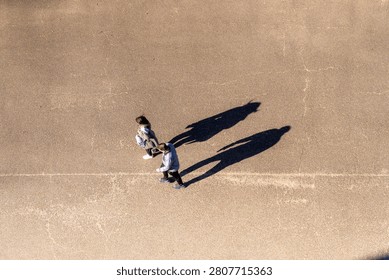 Looking straight down from above at two women in pale coats strolling close together over the bare textured pavement of the empty square of a public park in Japan in warm low winter light