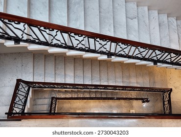 Looking straight down the center of multiple levels of a large, marble spiral staircase with a decorative handrail
