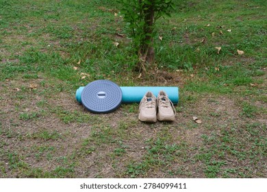 Looking straight down from an elevated position, a rolled blue yoga mat, a kinetic waist twister, and brown cream shoes are organized on a green park lawn.