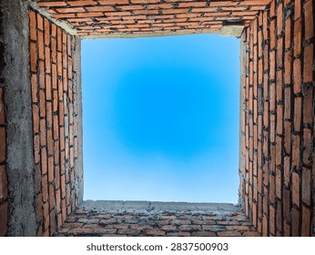 Looking straight upward from inside a rectangular vertical shaft framed by rough red masonry blocks reveals a vibrant azure sky overhead. The rustic clay construction shows weathered surfaces and uneven mortar joints surrounding the open aperture. Natural daylight illuminates the textured interior walls, creating a striking geometric perspective from deep within the hollow structure.