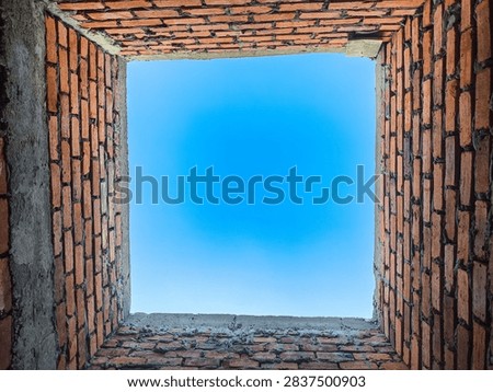 Looking straight upward from inside a rectangular vertical shaft framed by rough red masonry blocks reveals a vibrant azure sky overhead. The rustic clay construction shows weathered surfaces and uneven mortar joints surrounding the open aperture. Natural daylight illuminates the textured interior walls, creating a striking geometric perspective from deep within the hollow structure.