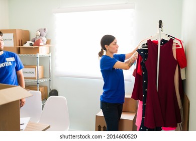 Looking Through The Clothes Rack At The Donation Center. Female Volunteer Looking For Clothing To Give To Someone In Need