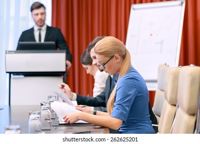 Looking through the documents. Closeup with selective focus on attractive blonde businesswoman in spectacles looking down to the papers while male business manager stands at the tribune on the