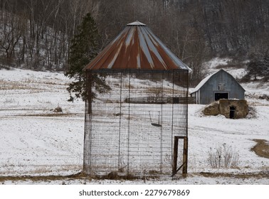 looking through an empty metal silo with a red roof and a grey barn in the distance on a snowy day in the country