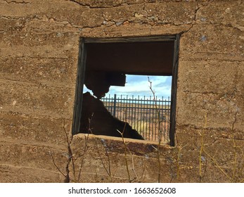 Looking through an opening where a window use to be of an old decaying and abandoned jail house in shades of brown with iron fencing, a desert field and a cloudy sky in the background.