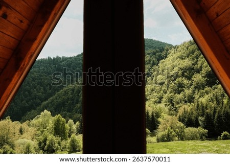 Looking through a triangular attic window toward green wooded hills. A-frame cabin in Tara National Park, Serbia. Peaceful nature and architectural charm combined.