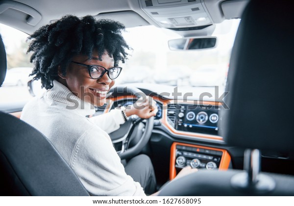 Looks back. Young african american woman sits inside
of new modern car.