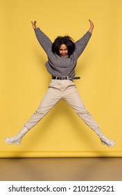 Looks Excited. Full-length Portrait Of Young Happy Dark Skinned Girl In Warm Knitted Sweater Jumping Isolated On Yellow Background. Concept Of Emotions, Facial Expression, Youth, Sales And Aspiration.