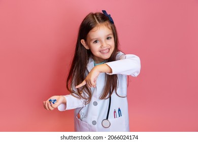 Looks Happy. Studio Shot Of Beautiful Little Girl, Child Playing Doctor Wearing White Lab Coat Isolated On Pink Background. Concept Of Childhood, Studying, Leisure Games. Health Care And Medicine.