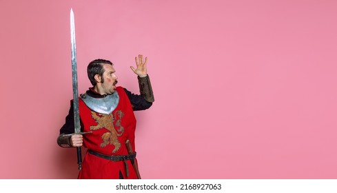 Looks Scared, Shocked. Portrait Of Medieval Warrior Or Knight Wearing Wearing Armor Clothing Holding Big Sword Isolated Over Pink Background. Comparison Of Eras, History, Funny Meme Emotions