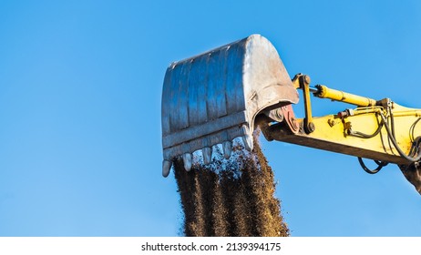 Loose soil falling down from excavator shovel lit by spring sunlight on a blue sky background. Close-up of yellow digger arm metal bucket when dumping brown dirt. Working hydraulic earth mover detail.