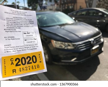 LOS ANGELES, April 7th, 2019: Close up of a new yellow DMV 2020 car registration sticker next to a blurry black Volkswagen and another car, parked on a residential street, in the background.