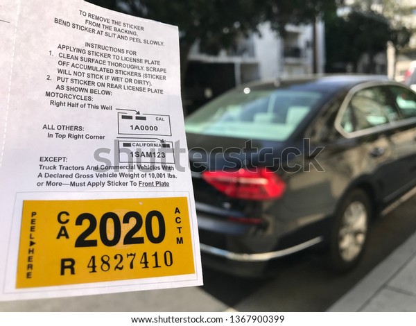 LOS ANGELES, April\
7th, 2019: Close up of a new yellow DMV 2020 car registration\
sticker next to a blurry black Volkswagen, parked on a residential\
street, in the background.