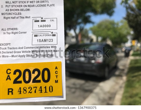 LOS ANGELES,\
April 8th, 2019: Close up of a new yellow DMV 2020 car registration\
tag next to the back of a black Prius, parked on a quiet\
residential street, in the\
background.