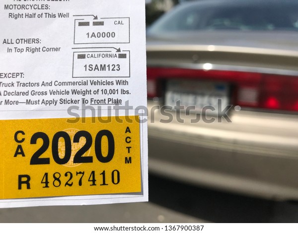 LOS ANGELES, April 8th, 2019: Close up of a\
new yellow DMV 2020 car registration sticker next to the license\
plate of a blurry brown older Buick, parked on a residential\
street, in the background.