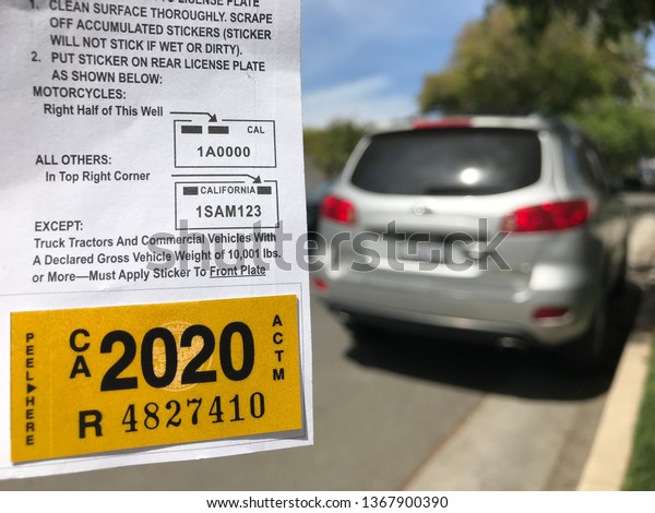 LOS ANGELES, April\
8th, 2019: Close up of a new yellow DMV 2020 car registration\
sticker next to a gray car, parked on a quiet residential street,\
in the background.