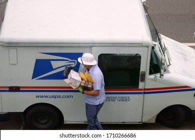 Los Angeles, CA / USA - April 20, 2020: A United States Postal Service USPS Mailman Wears A Mask And Gloves While Carrying A Load Of Parcels From A Mail Truck During The COVID-19 Coronavirus Pandemic.