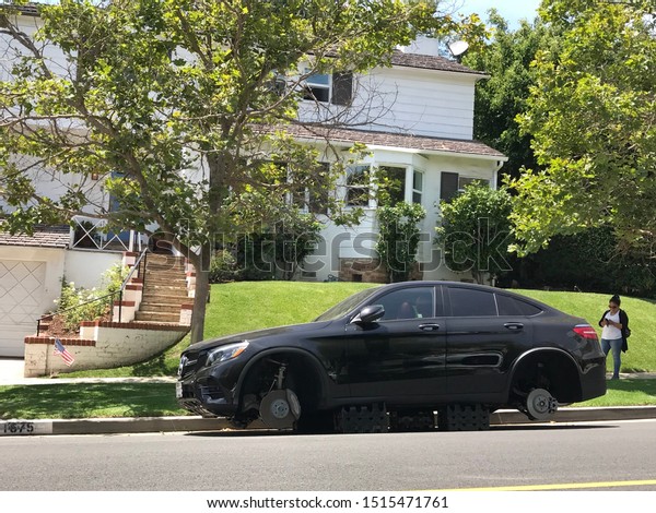 LOS ANGELES, June 26th, 2019: Woman with cell phone\
standing next to a black luxury Mercedes Benz car, stripped of its\
tires and standing on plastic crates next to an American flag, in\
an upscale 
