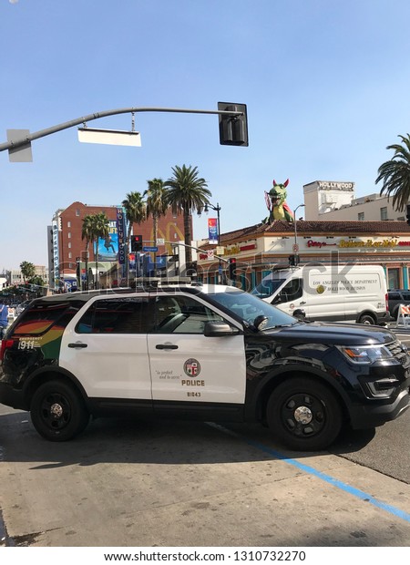 LOS ANGELES, Nov 10th, 2018: A police cruiser\
and a white Hollywood Division van stand across closed off\
Hollywood Boulevard and Highland Ave, with colorful Hollywood signs\
and palms in the background