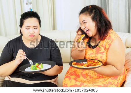 Lose weight concept. Unhappy fat woman eating salad while teasing by her sister eating pizza with mocking expression