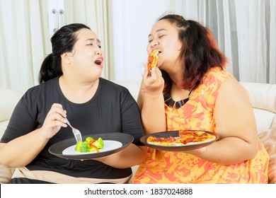 Lose Weight Concept. Young Obesity Woman Eating Pizza While Teasing Her Fat Sister Eating Salad In The Living Room. Shot At Home