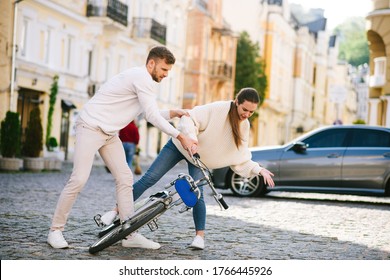 Lost Balance. Man In White Sweater And Jeans Picking Up A Fallen Bicycle With A Young Injured Woman