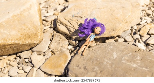 A lost doll with purple hair rests on large rocks by a riverbank under bright sunlight during a warm summer day. Dangerous children's game on the beach - Powered by Shutterstock