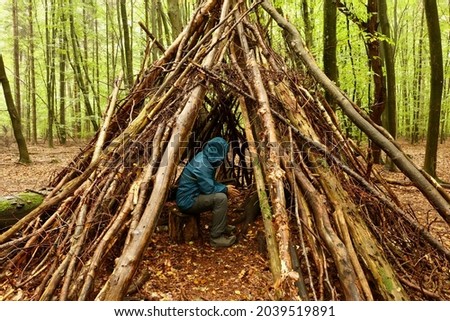 A lost hiker takes refuge in a makeshift shelter constructed of branches leaned against the trunk of a tree in a forest in northern Germany